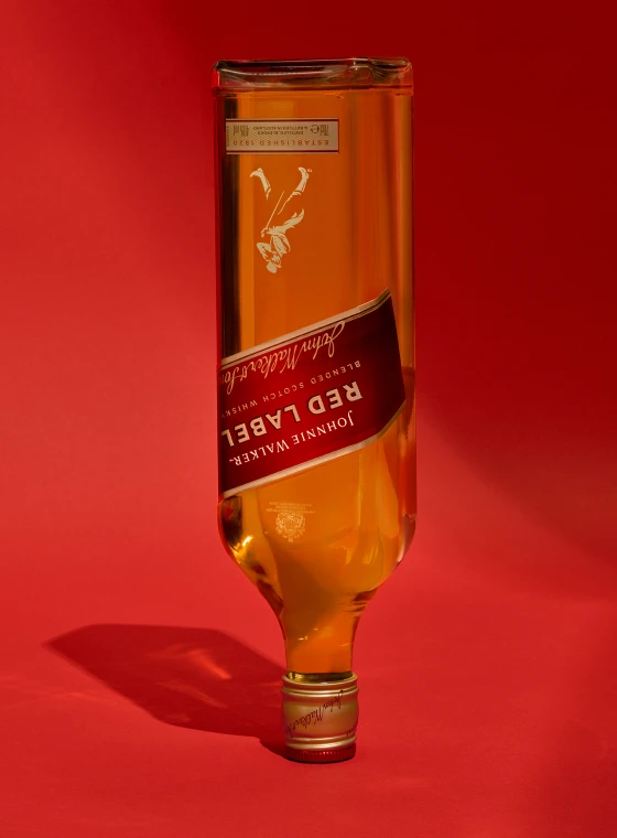 Пляшка шотландського віскі Johnnie Walker Red Label перевернута на яскраво-червоному тлі, відкидаючи тінь. Ярлик і логотип добре видно на передній частині.