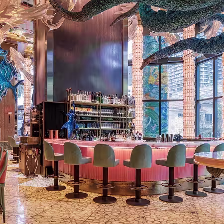 A modern, stylish bar with pink lighting, green swivel chairs, a curved marble counter, tall shelves of bottles, and large windows. Artistic ceiling decorations and sculptural elements add a whimsical touch.