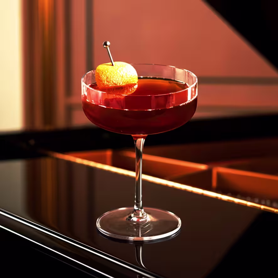 A deep red cocktail garnished with an orange peel sits in a coupe glass on a glossy black surface, reflecting warm, ambient lighting.
