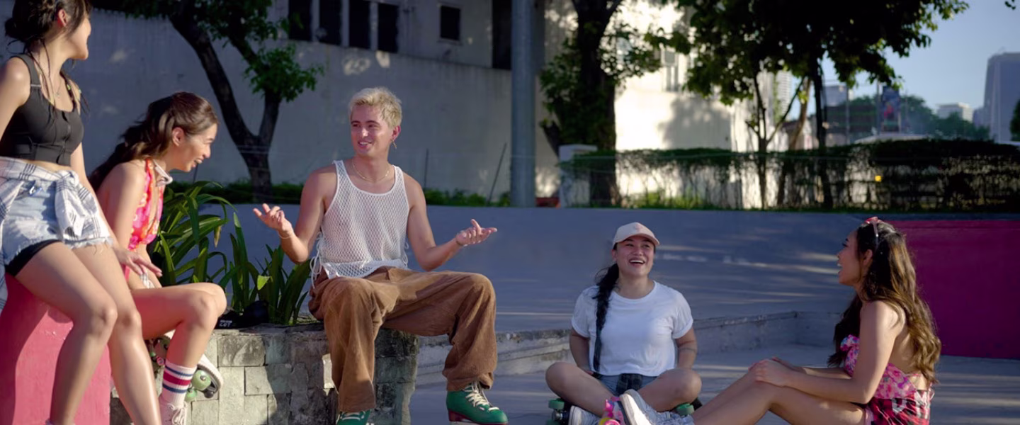 Five young people sit and laugh together outdoors at a skate park; some are wearing casual summer clothes and roller skates, enjoying a sunny day with trees and city buildings in the background.