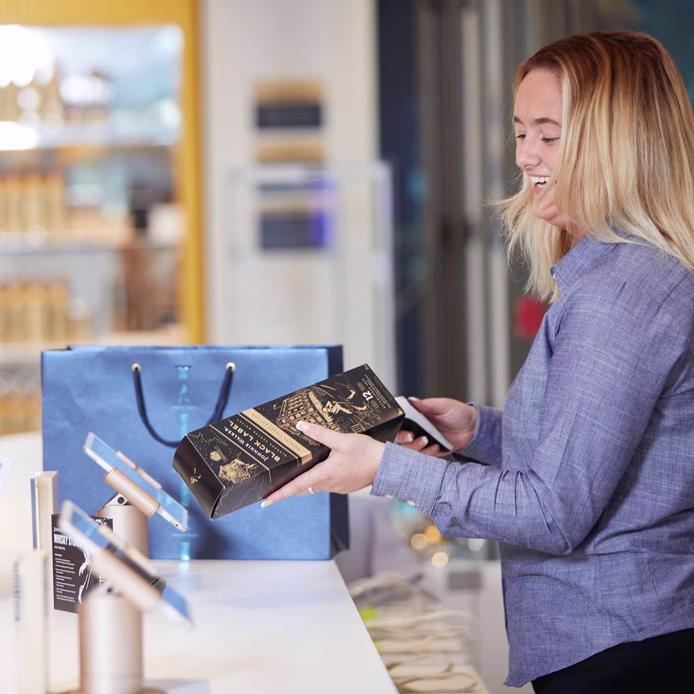 Smiling woman at a store counter holding a black and gold box next to a blue shopping bag.