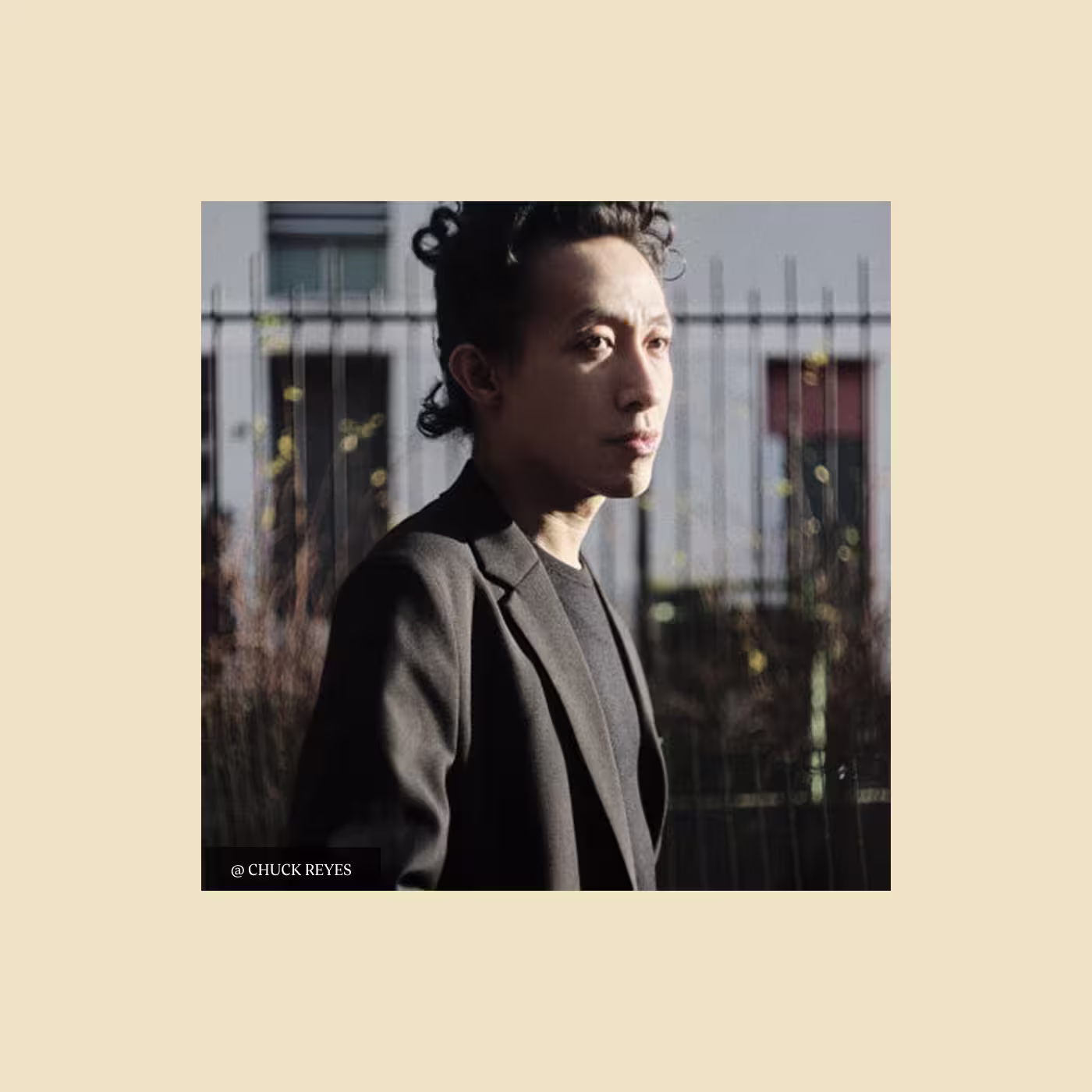 A person with short, curly dark hair wearing a black blazer and shirt stands outdoors in sunlight, looking to the side. A blurred fence and building are in the background. Photo credit: @CHUCK REYES.