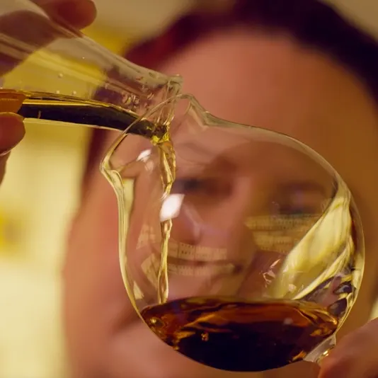 Person pouring liquid from a vial into a spherical glass container, creating ripples. The focus is on the liquid and glass, with the person blurred in the background. Warm lighting creates a cozy atmosphere.