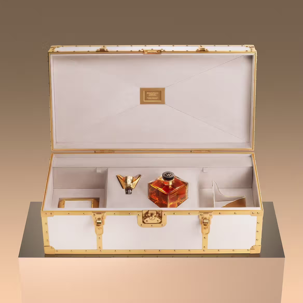 Một chiếc hộp sang trọng, viền kem và vàng với nội thất nhung, mở ra sẽ thấy một chai thủy tinh trang trí, một chiếc phễu màu vàng và các ngăn đựng phụ kiện, được trưng bày trên bề mặt phản chiếu.