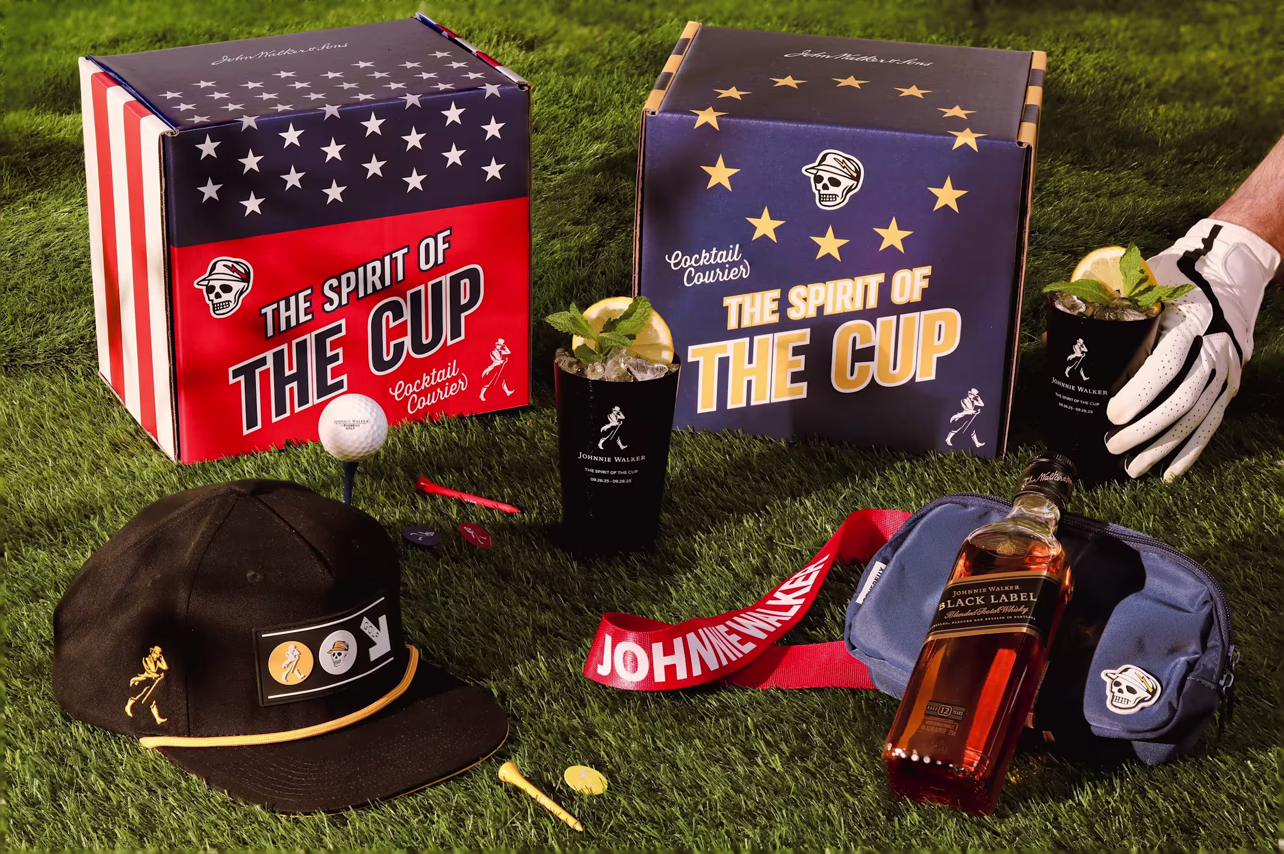 Boxes labeled The Spirit of the Cup, golf-themed items, two cocktails, a bottle of Johnnie Walker Black Label, a golf glove, hat, and golf balls arranged on green grass.
