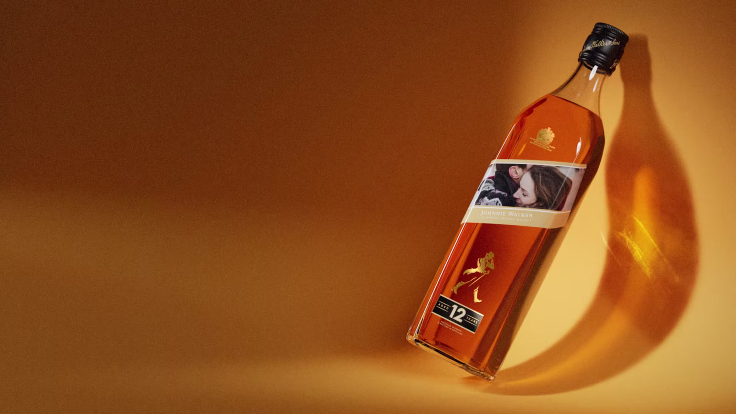 Une bouteille de whisky Johnnie Walker Black Label 12 ans d'âge repose sur une surface orange, projetant une ombre. L'étiquette présente une photo de deux personnes enlacées.