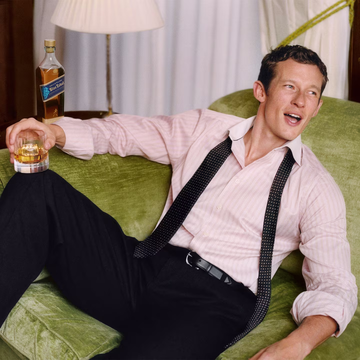 Callum Turner in dress shirt and tie sits on green sofa, holding a glass of whiskey, looking off to the side.