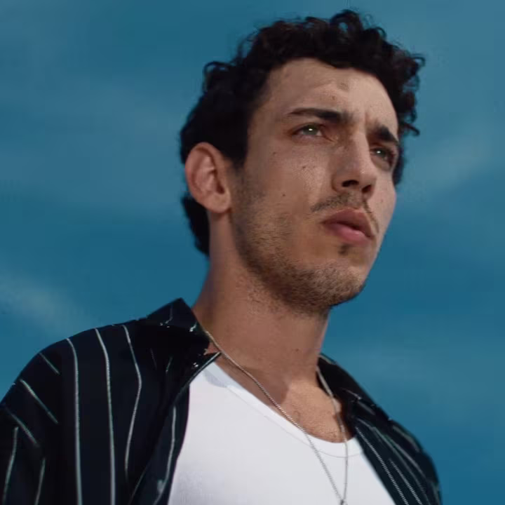 Man with short curly hair and striped shirt looks into the distance against a blue sky.