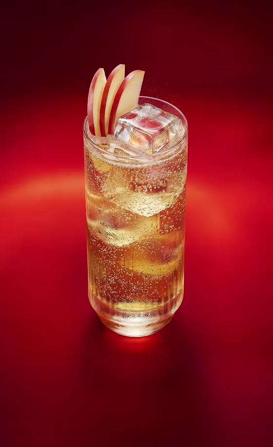 Una bebida espumosa con hielo y rodajas de manzana como decoración en un vaso transparente, sobre un fondo rojo.