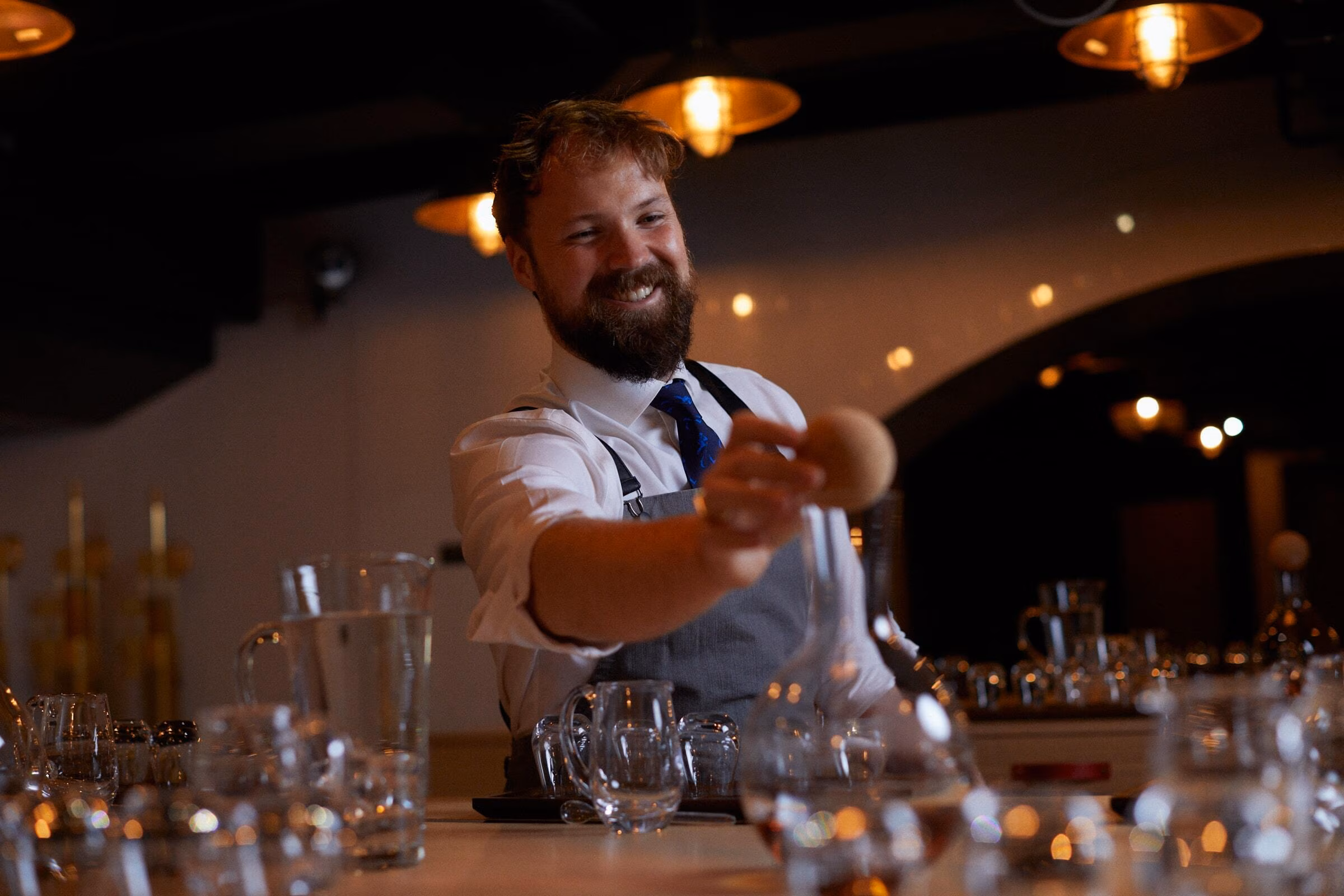 A bearded bartender smiles while placing an ice sphere into a glass at a dimly lit bar.