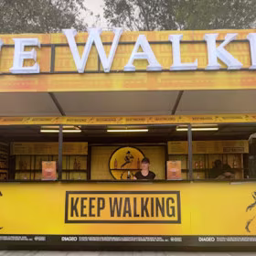 Un puesto de bebidas al aire libre de color amarillo brillante y negro con un letrero de "Keep Walking" al frente y un vendedor detrás del mostrador. La parte superior del puesto luce letras grandes con el nombre de la marca. Se ven árboles al fondo.