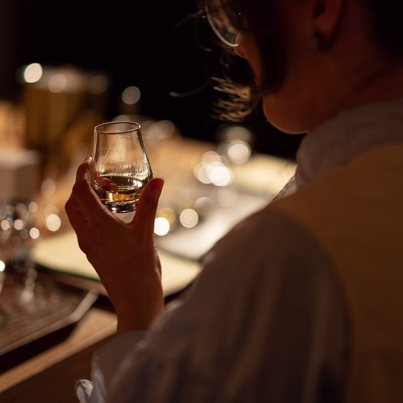 A person holding a glass of whiskey, seen from behind, in a softly lit setting.