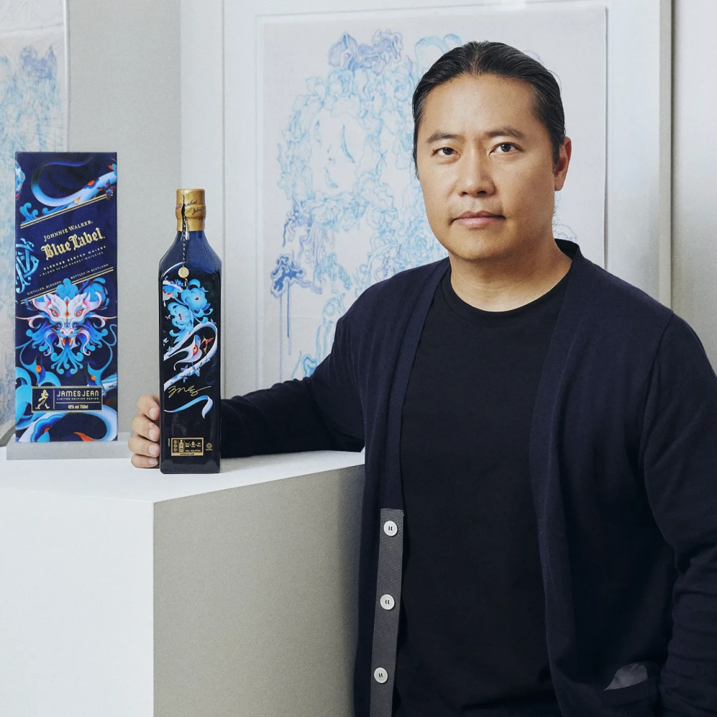 James Jean steht neben einer Flasche Johnnie Walker Blue Label. Die Flasche und die Schachtel sind mit farbenfrohen, kunstvollen Motiven versehen. Die Person trägt eine dunkle Strickjacke und steht vor einem gerahmten Kunstwerk an einer weißen Wand.