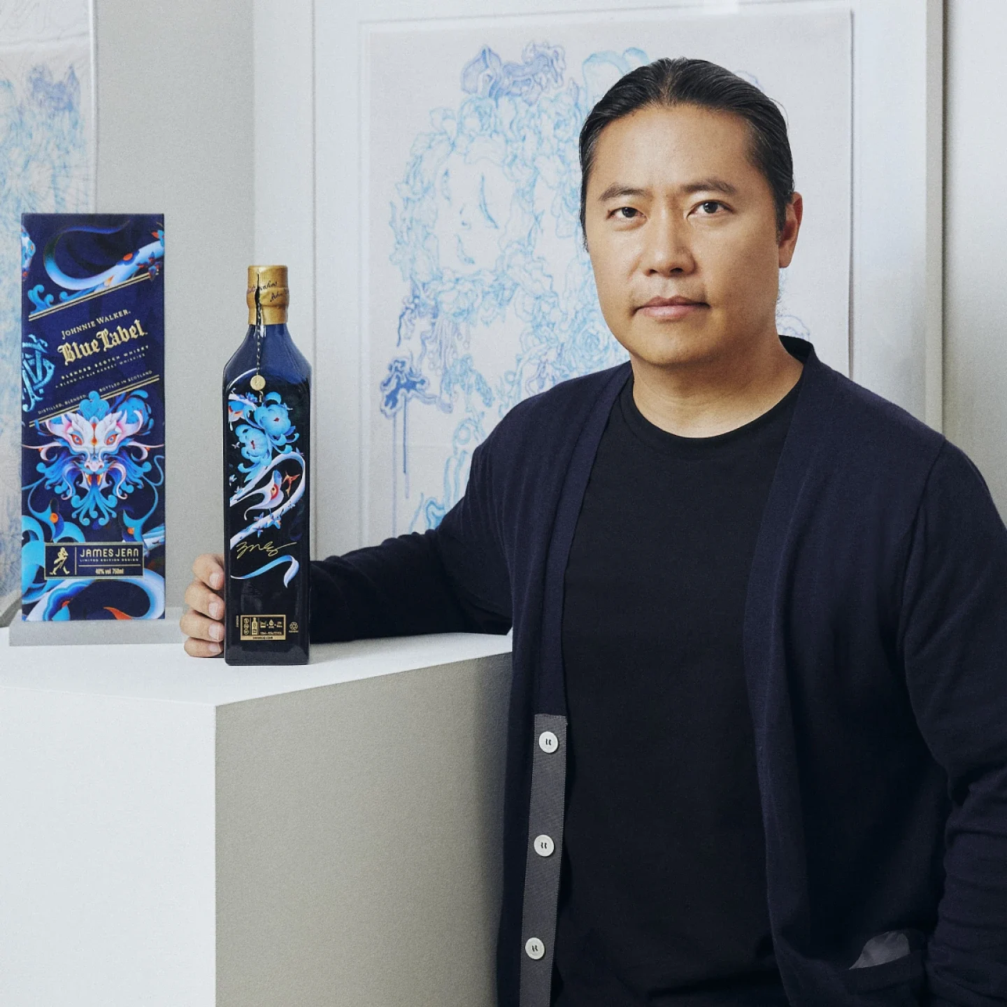 Un homme aux longs cheveux noirs se tient à côté d'une bouteille Johnnie Walker Blue Label en édition limitée présentant des illustrations bleues complexes, avec un emballage assorti et des illustrations bleues encadrées en arrière-plan.