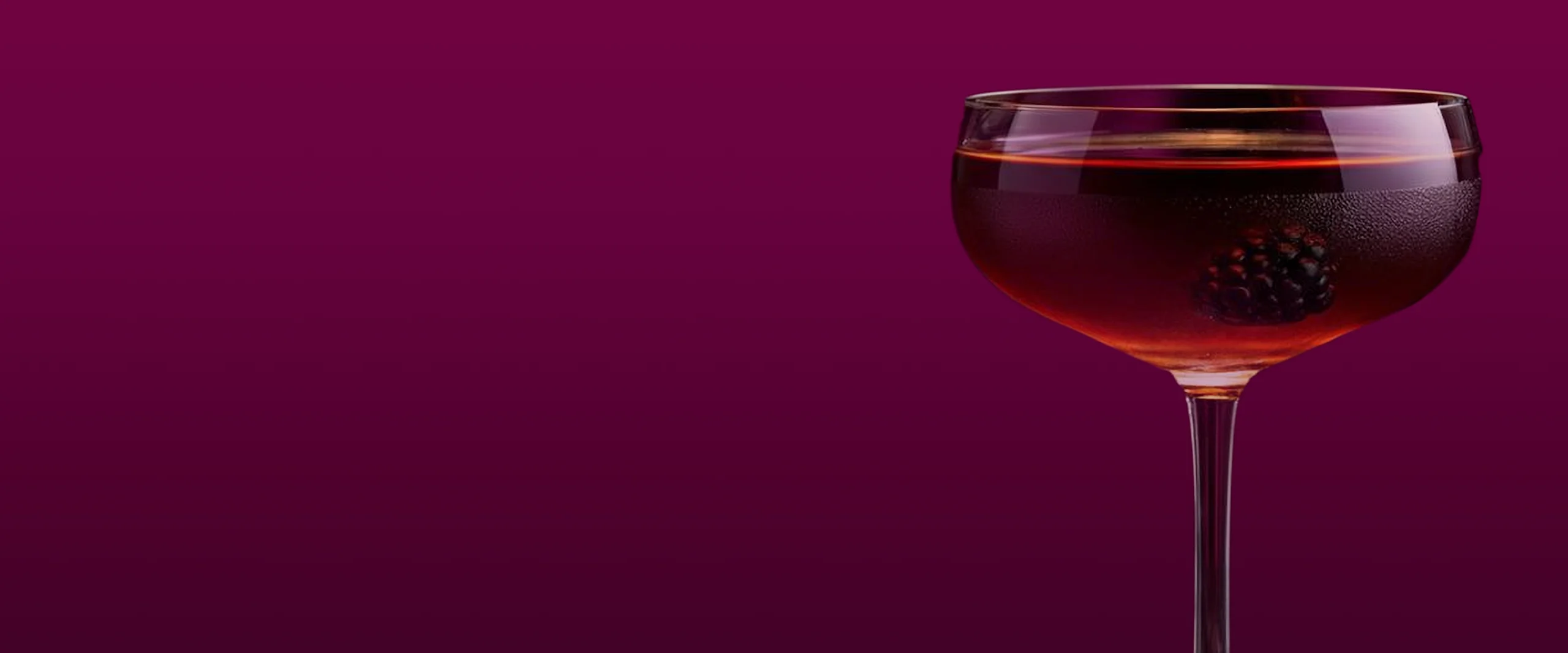A stemmed glass filled with a dark red liquid, garnished with a blackberry, on a soft, deep purple background.
