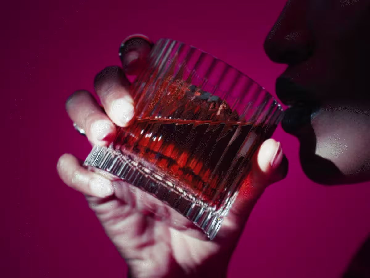 Een persoon met donkere lippenstift houdt een glas amberkleurige vloeistof dicht bij zijn lippen tegen een levendige roze achtergrond. Het drankje lijkt in een getextureerd tumblerglas te zitten, en de persoon heeft gemanicuurde nagels en draagt ​​ringen.