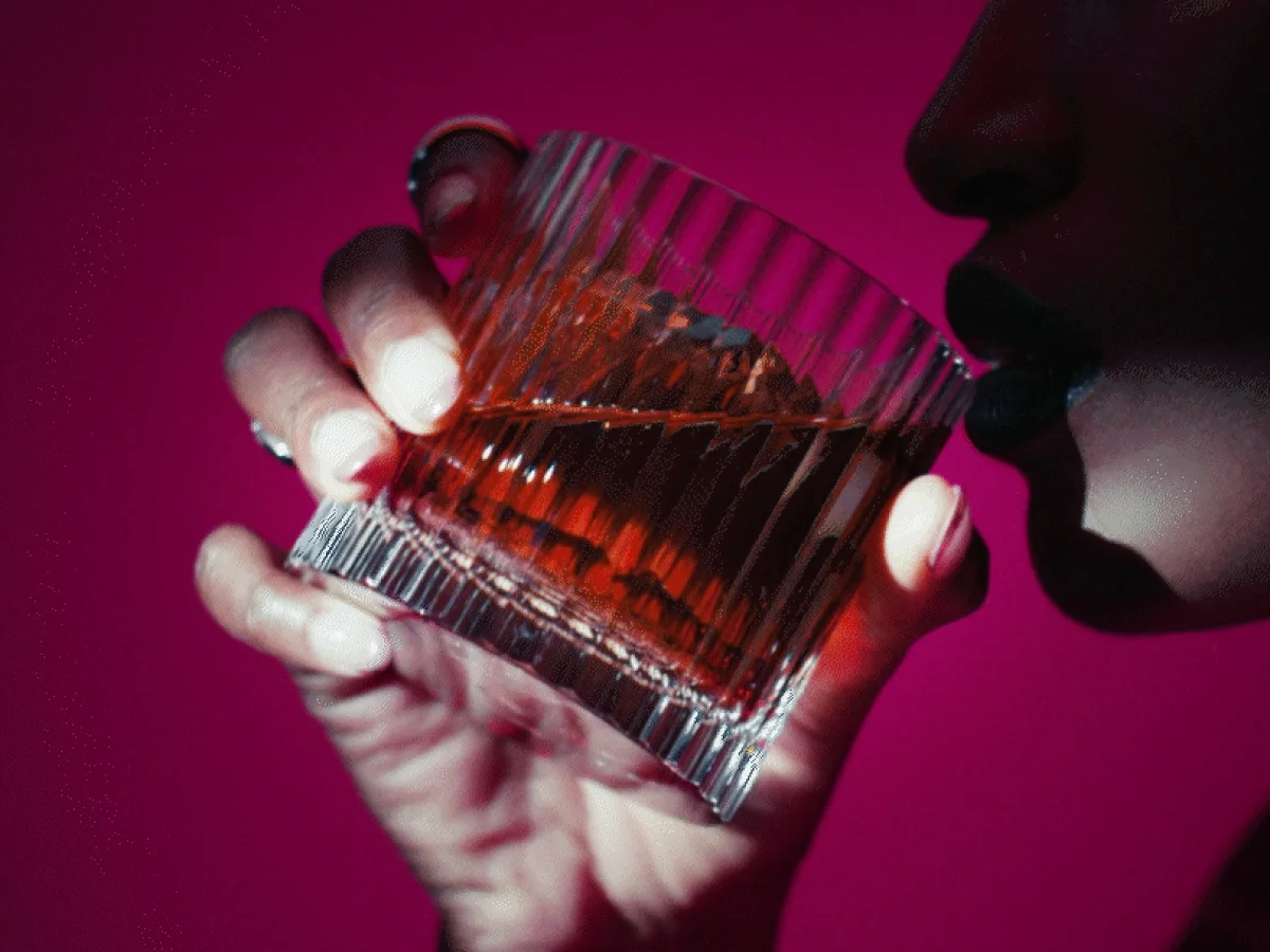 A person holding a glass of amber-colored Black Ruby whisky, against a deep pink background. The glass is faceted, and the person is about to take a sip. The lighting casts dramatic shadows on their hand and face.