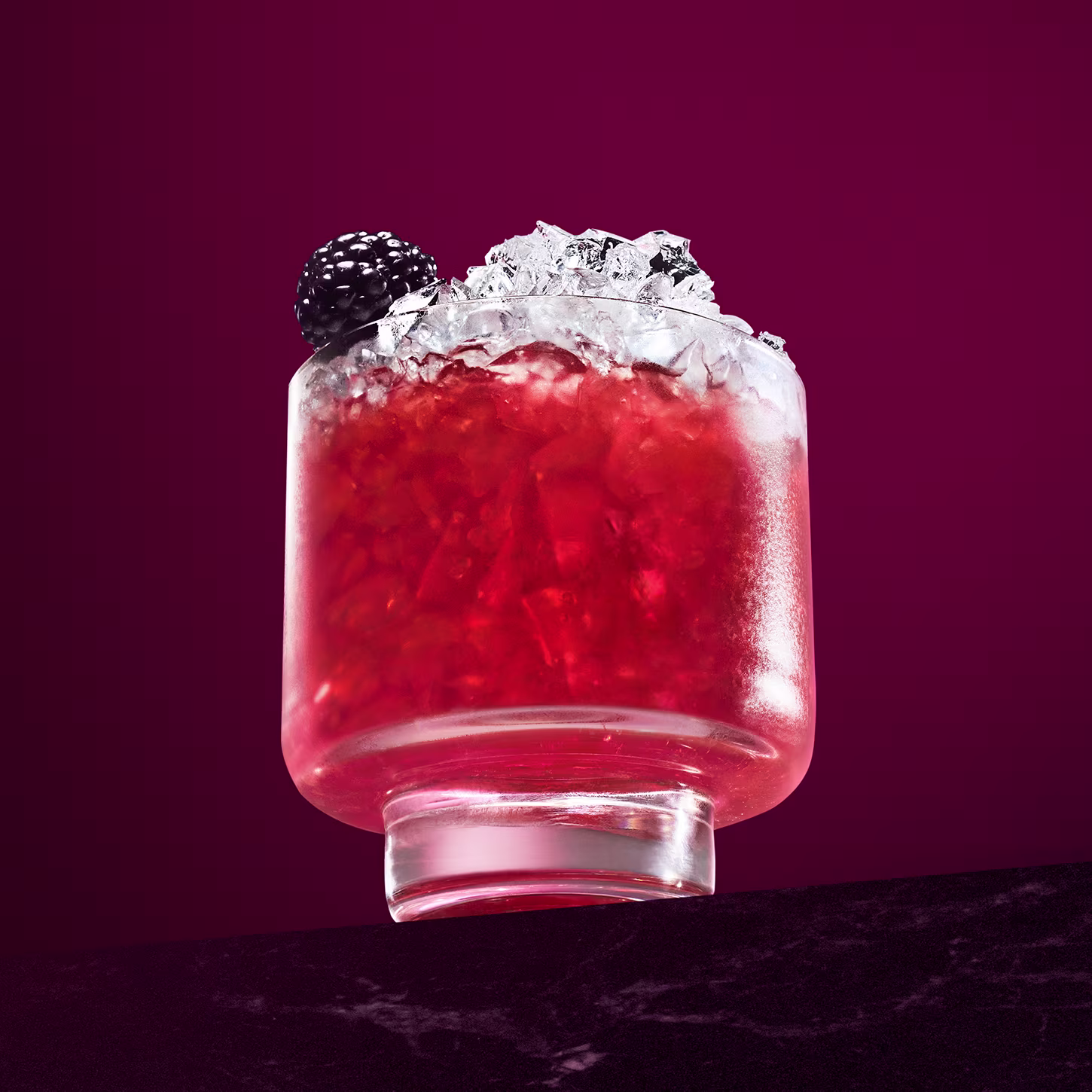 Ein leuchtend roter Cocktail in einem klaren Glas, mit zerstoßenem Eis und einer Brombeere garniert. Der Hintergrund ist tiefmagentafarben und verleiht dem Getränk einen besonderen Kontrast.