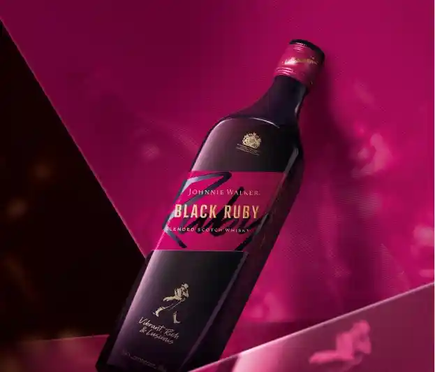 Una botella de whisky escocés Johnnie Walker Black Ruby Blended se encuentra sobre una superficie rosa. La etiqueta de la botella presenta texto dorado y el logotipo de una figura caminando.