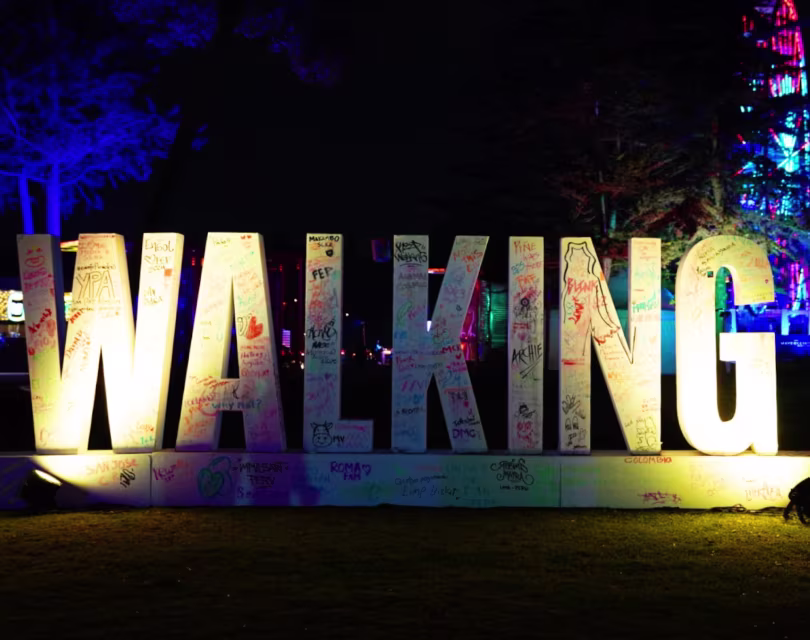 Las letras iluminadas que forman la palabra WALKING, cubiertas de coloridos grafitis, se exhiben al aire libre por la noche. Cerca de allí, una estatua de un hombre con sombrero de copa y traje realza la escena. 