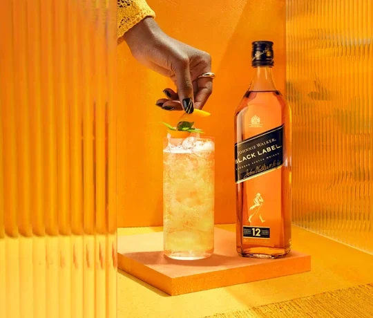The Crisp Citrus Johnnie & Lemon. A hand garnishes a tall iced cocktail with a citrus twist and mint, next to a bottle of Johnnie Walker Black Label whiskey. The background is a vibrant orange with textured panels, enhancing the warm tones of the scene.