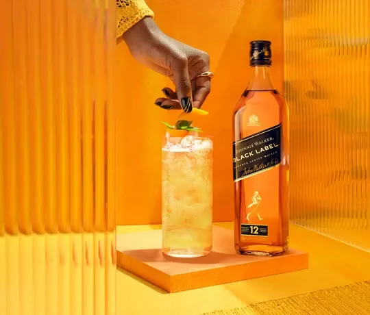 Ręka zdobi wysoką szklankę lodowatego koktajlu z cytrusowym akcentem i listkiem mięty. Stoi obok butelki Johnnie Walker Black Label na jasnopomarańczowym tle.
