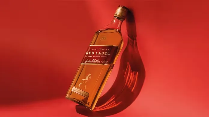  Una botella de whisky Johnnie Walker Red Label reposa inclinada sobre una superficie roja vibrante, proyectando una sombra. La etiqueta y el logotipo de la firma son claramente visibles en la botella.