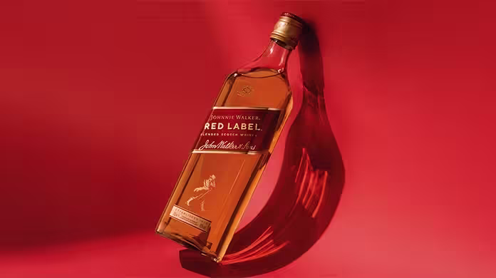  Uma garrafa de uísque Johnnie Walker Red Label repousa inclinada sobre uma superfície vermelha vibrante, projetando uma sombra. O rótulo e o logotipo da assinatura são claramente visíveis na garrafa.