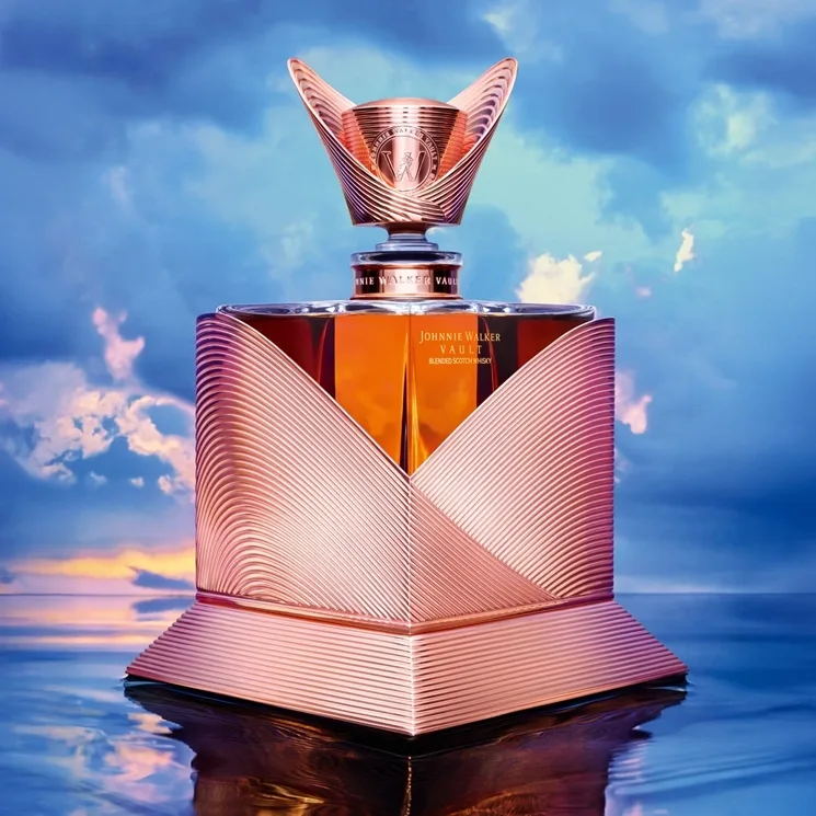 A luxurious, gold-toned Johnnie Walker bottle with intricate geometric patterns sits on a reflective surface, set against a vibrant sky with clouds and soft sunlight.