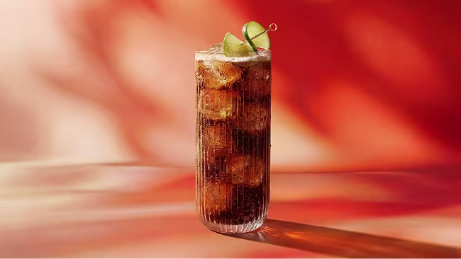 A tall glass of iced cola with lime wedges on top, set against an orange and red background.