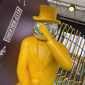 Una estatua de una persona con traje amarillo brillante y sombrero de copa, con la cabeza en forma de bola de discoteca, se encuentra frente a un cartel de "Sigue caminando". La figura sostiene un bastón amarillo y gesticula con una mano, sobre 