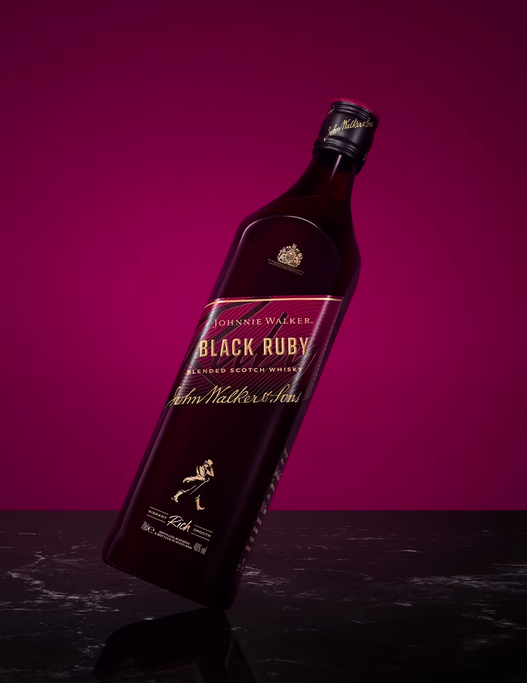 A bottle of Johnnie Walker Black Ruby blended Scotch whiskey is tilted against a dark red background. The label features gold text and details. The bottle is placed on a dark, reflective surface.