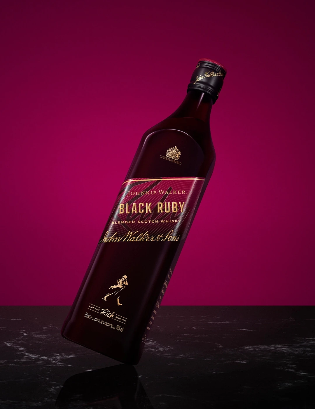 A bottle of Johnnie Walker Black Ruby blended Scotch whiskey is tilted against a dark red background. The label features gold text and details. The bottle is placed on a dark, reflective surface.