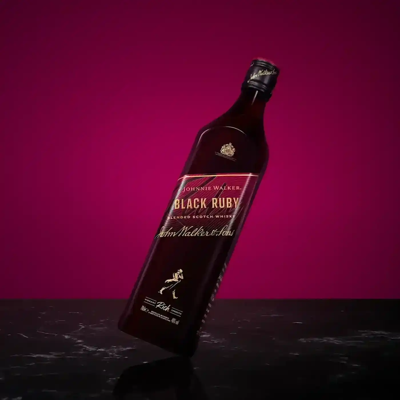 Ένα γερμένο μπουκάλι Johnnie Walker Black Ruby σε ένα πλούσιο μπορντό φόντο. Το μπουκάλι έχει χρυσά γράμματα και το εμβληματικό λογότυπο του striding man. Στηρίζεται σε μια ανακλαστική σκοτεινή επιφάνεια.