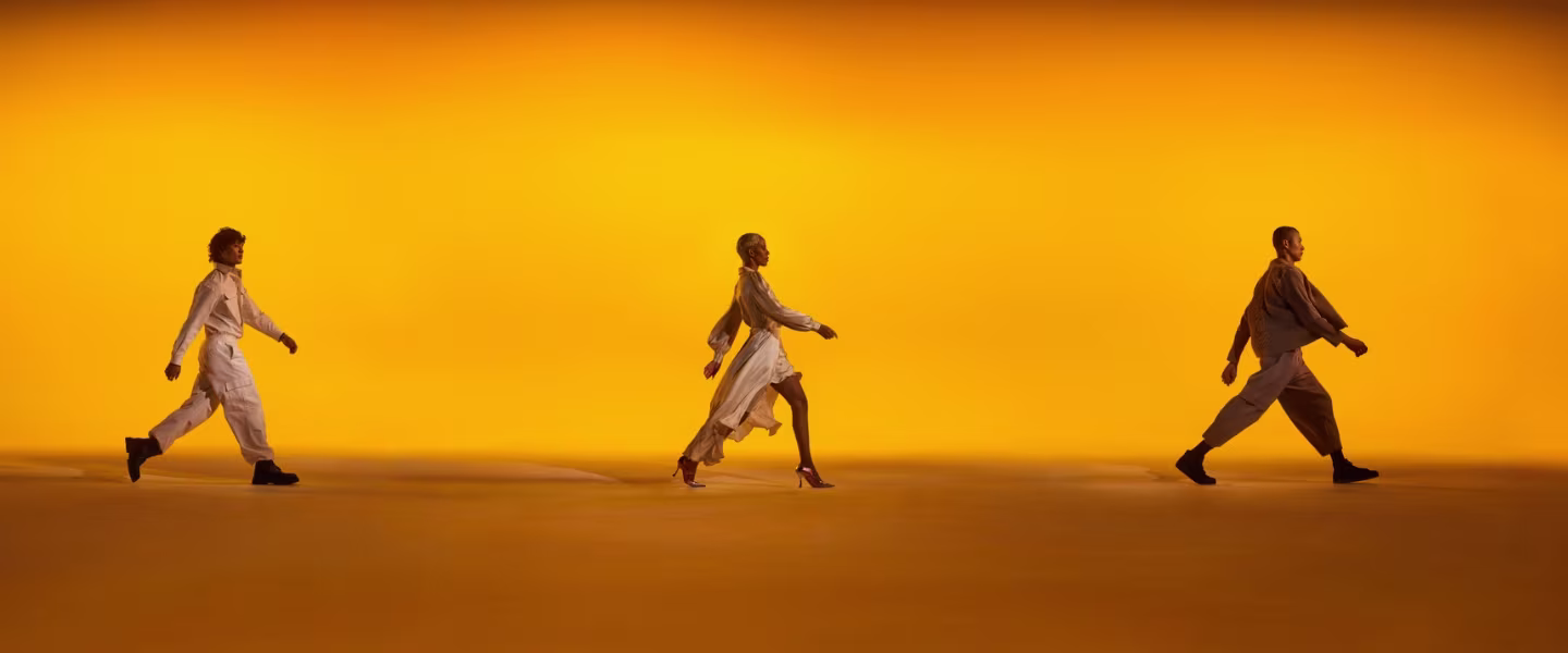 Drie personen lopen synchroon van links naar rechts over een levendige oranje achtergrond. Elke persoon vertoont een eigen loophouding en ze dragen stijlvolle, neutrale outfits. 