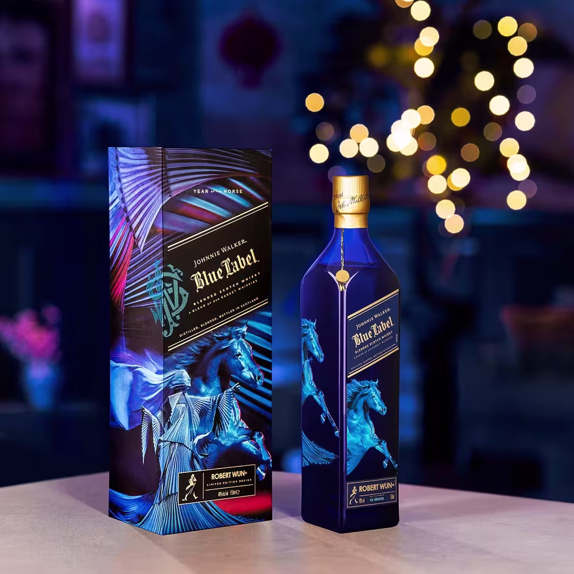 Ένα μπουκάλι ουίσκι Johnnie Walker Blue Label περιορισμένης έκδοσης και το κουτί του, και τα δύο με έργα τέχνης από το μπλε άλογο, εκτίθενται σε ένα τραπέζι με θολά ζεστά φώτα και ένα άνετο δωμάτιο στο βάθος.