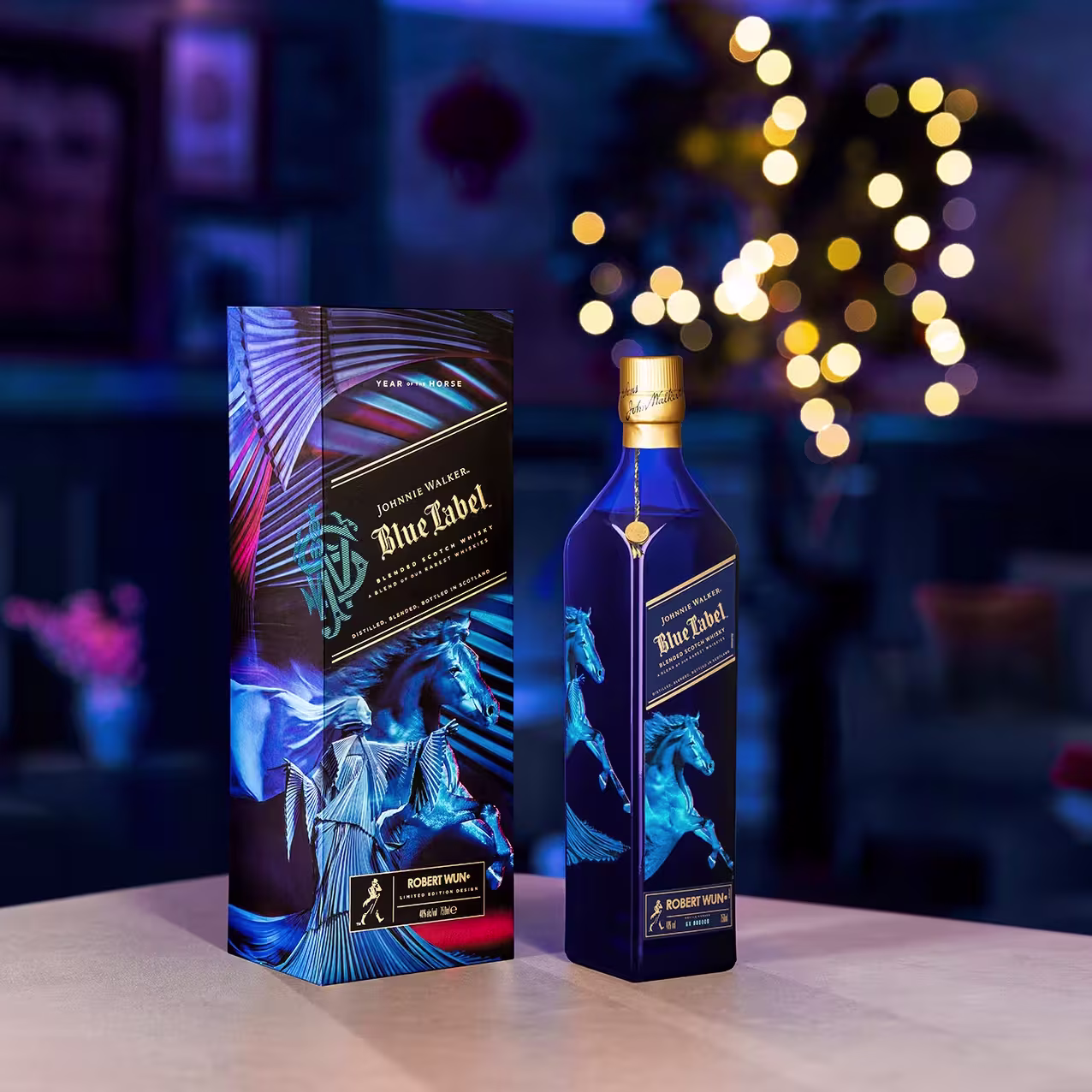 A bottle of Johnnie Walker Blue Label whisky stands next to its decorative box on a table, with a blurred, softly lit living room and string lights in the background.