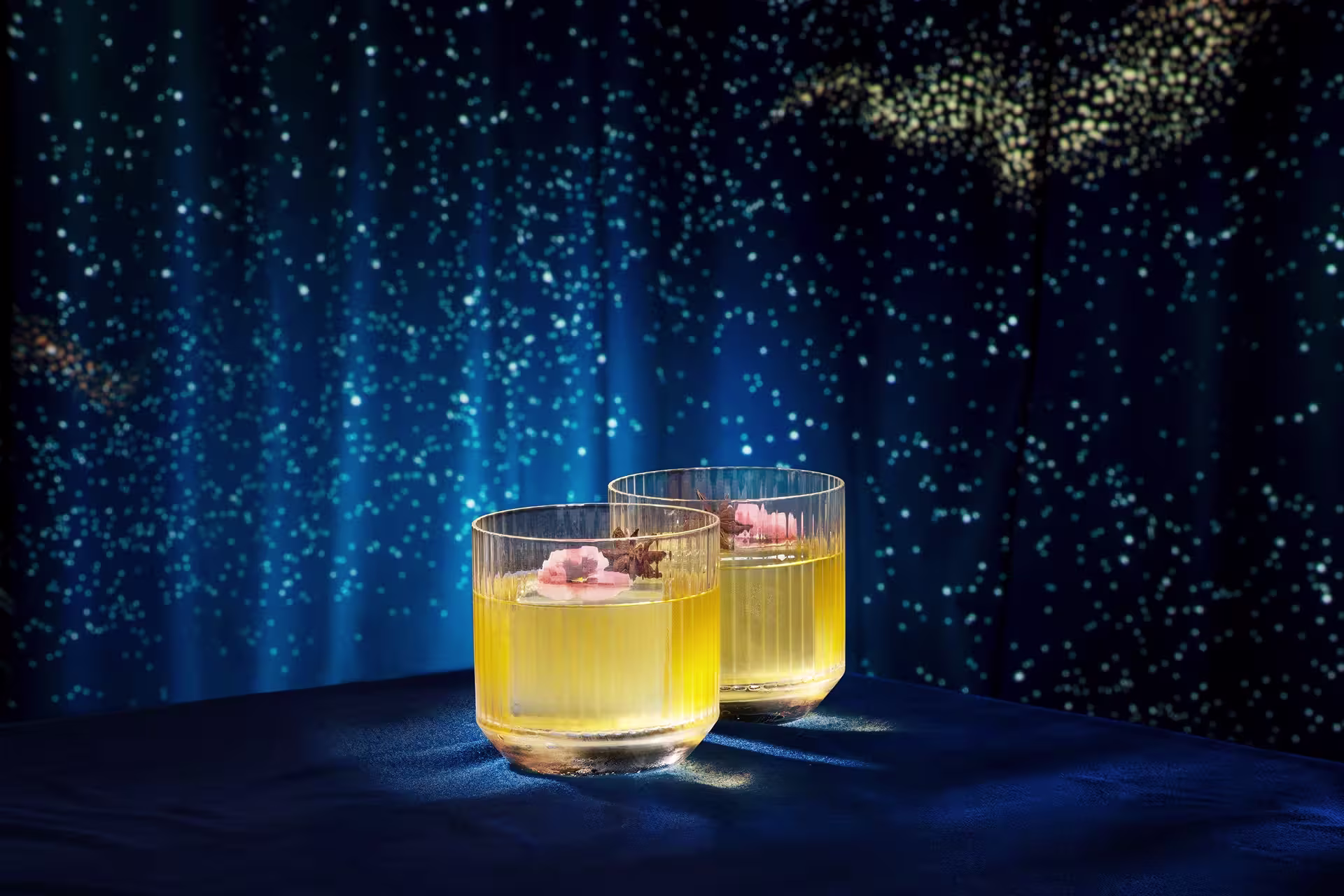 Two glasses of yellow cocktail garnished with pink flowers, placed on a blue surface with a starry night sky backdrop.