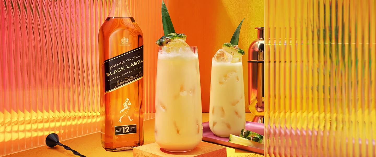A bottle of Johnnie Walker Black Label sits next to two pineapple-laced cocktails filled with ice. The setting is vibrant, with warm shades of orange and yellow, accented by a pineapple slice on a pink serving tray.