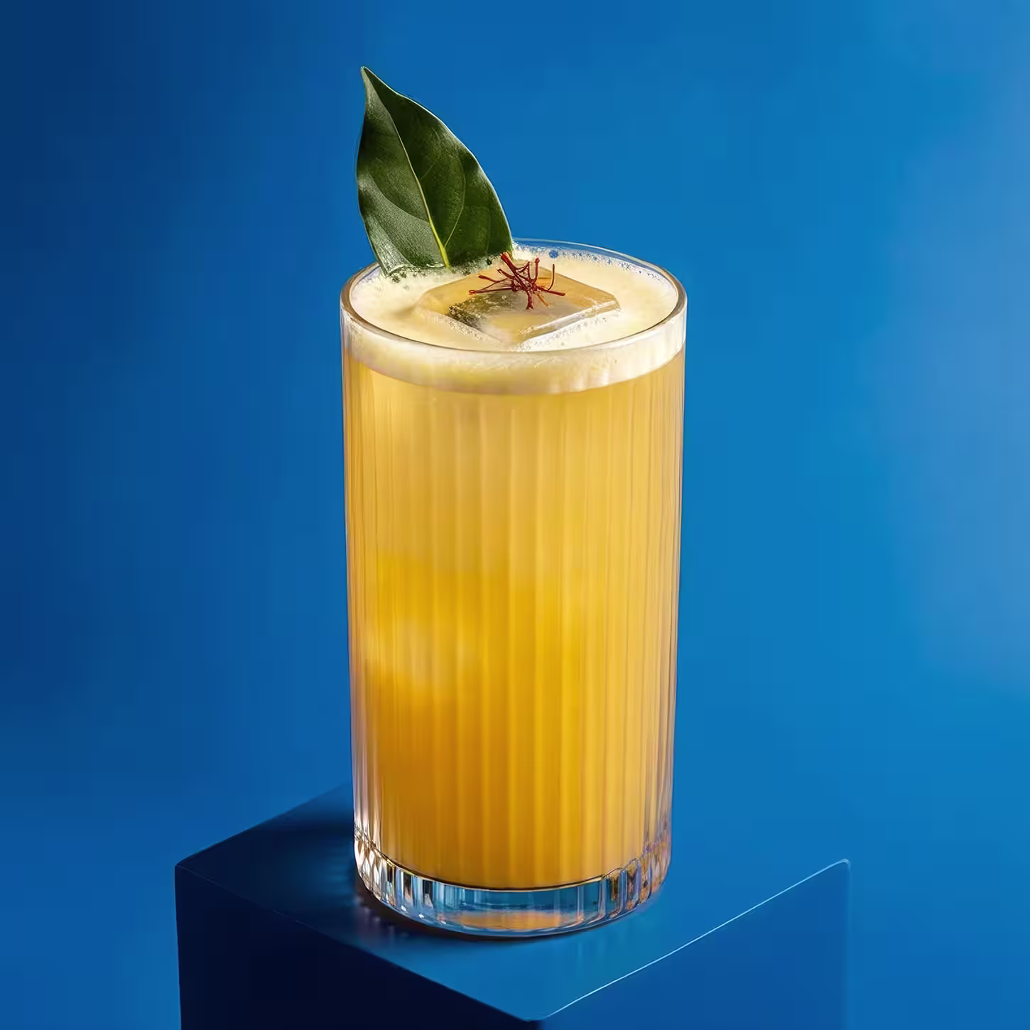 A tall glass filled with a bright yellow-orange drink, topped with a large ice cube, a bay leaf, and a few saffron strands, set against a solid blue background.