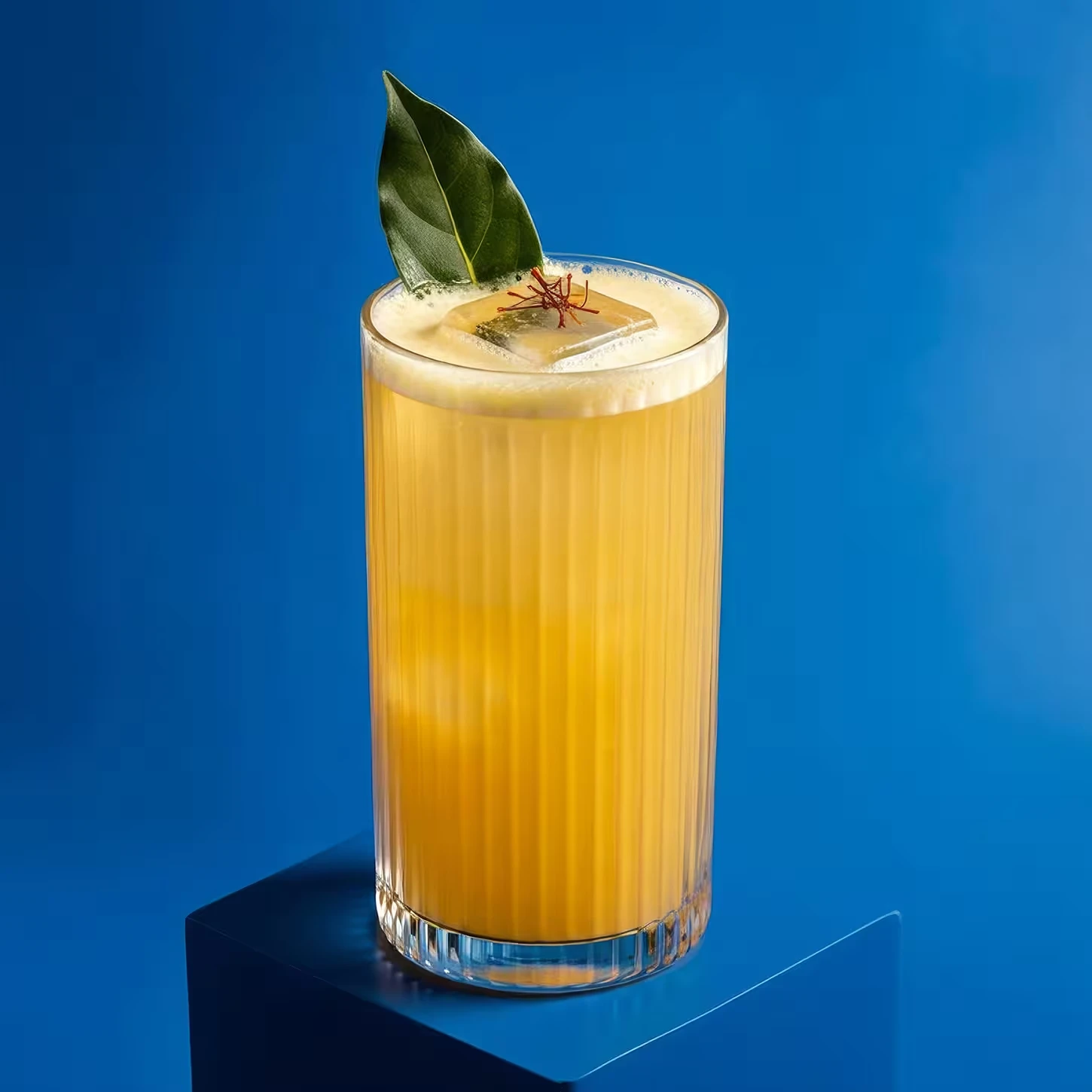 A tall glass filled with a bright yellow-orange drink, topped with a large ice cube, a bay leaf, and a few saffron strands, set against a solid blue background.