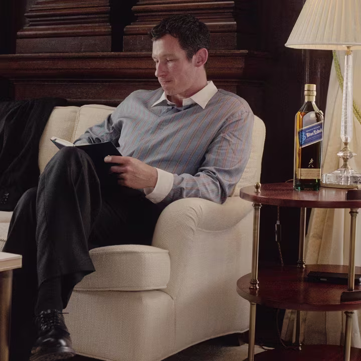 Callum Turner sitting on a white chair reading a notebook, with a bottle of whisky on a side table nearby.