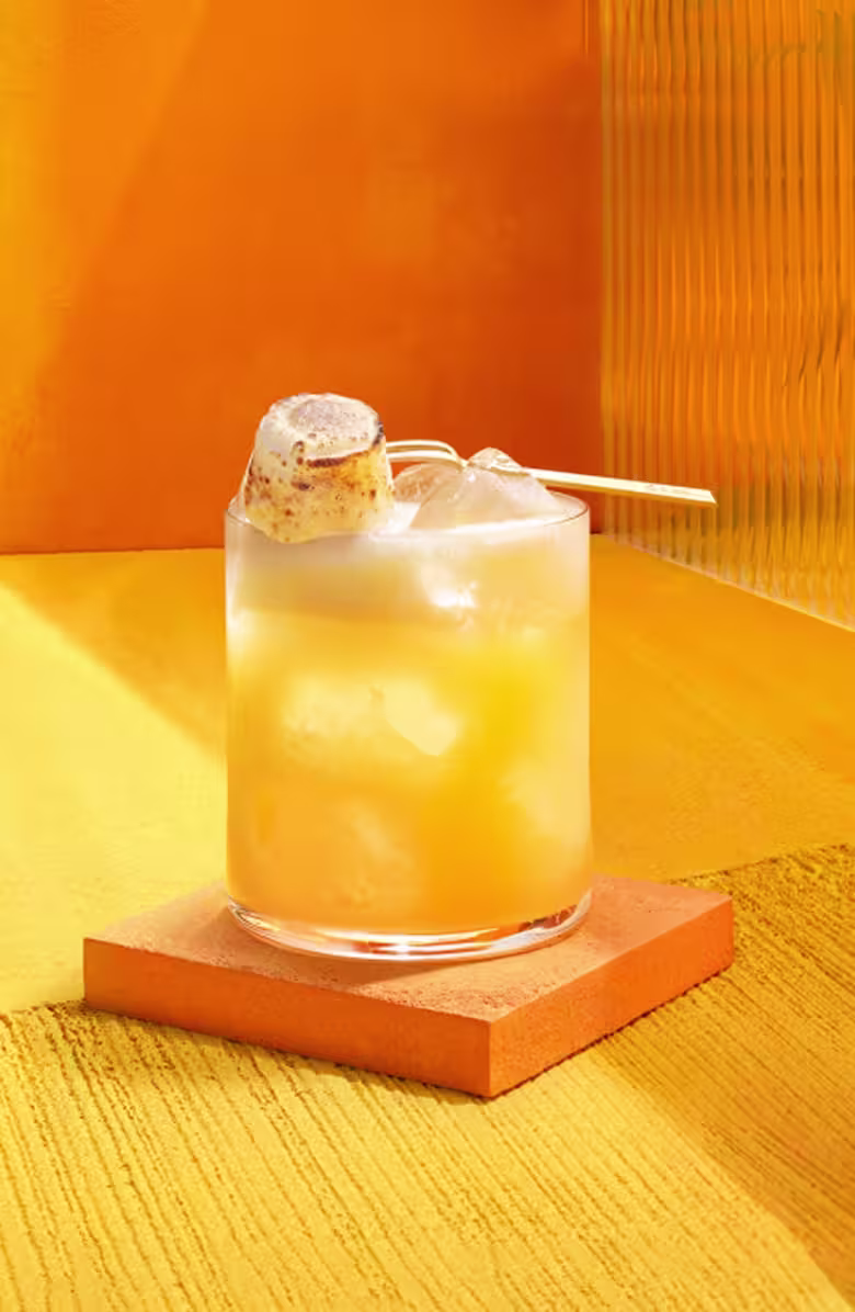 The Smooth & Floral Old Fashioned. A glass of iced orange beverage sits on an orange coaster. The drink is garnished with a toasted marshmallow on a skewer. The background is a gradient of orange hues with textured surfaces.