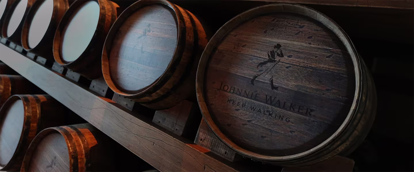 Редица дървени бъчви с логото „Johnnie Walker Keep Walking“ върху тях, подредени хоризонтално върху дървена поставка. Осветлението е слабо, подчертавайки наситените кафяви тонове на дървото.