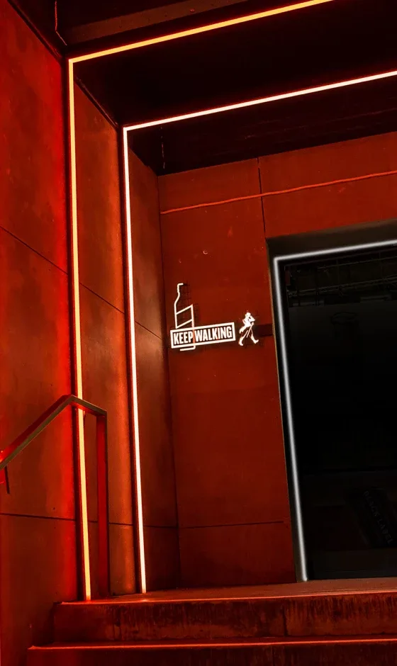 Een zwak verlichte trap met rode verlichting leidt naar boven. Op een bord bovenaan staat het silhouet van een lopend persoon en de woorden KEEP WALKING. De muren zijn diep oranjebruin en versterken de warme gloed.
