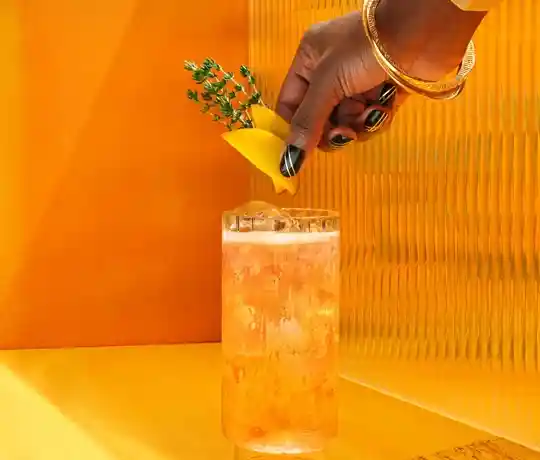 Een hand met zwarte en gouden nagels, versierd met gouden armbanden, versiert een groot glas ijskoude sinaasappeldrank met een citroenschil en kruiden. De achtergrond is levendig geel met een gestructureerd oppervlak.