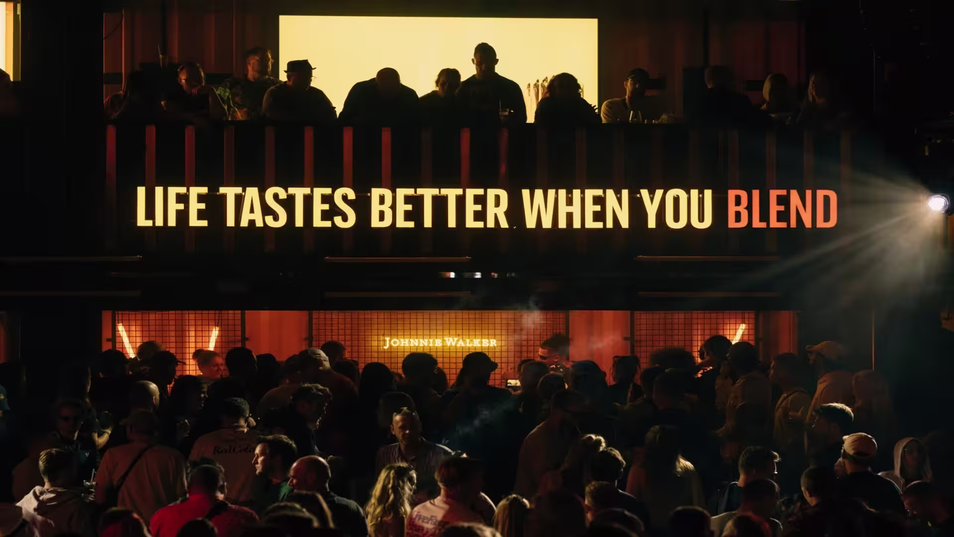 Una gran multitud se reúne por la noche bajo un cartel brillantemente iluminado que dice “La vida sabe mejor cuando te mezclas”, con siluetas de personas en un nivel superior por encima de la multitud.