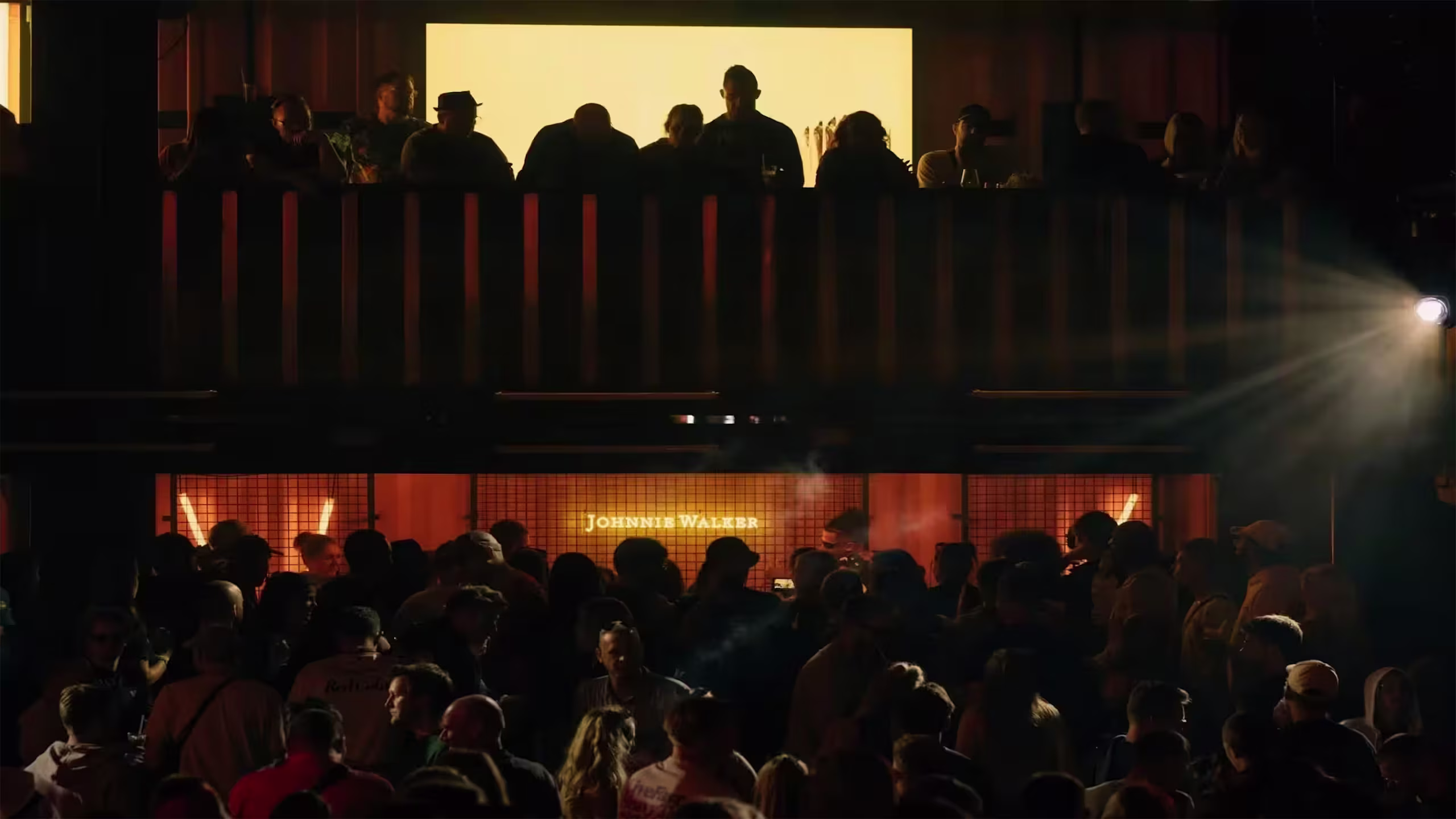 Eine große Menschenmenge versammelt sich nachts unter einem hell erleuchteten Schild mit der Aufschrift „Das Leben schmeckt besser, wenn man sich vermischt“, wobei auf einer oberen Ebene über der Menge Menschen als Silhouetten zu erkennen sind.