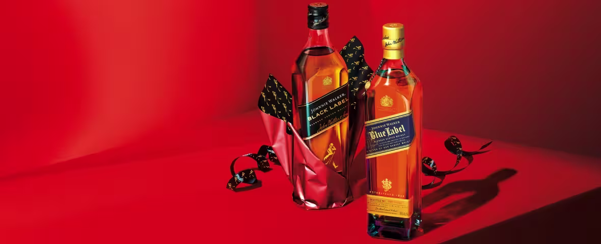 Two bottles of Johnnie Walker whisky, one Black Label partially wrapped in red paper and one Blue Label, standing on a red surface with a ribbon and red background.