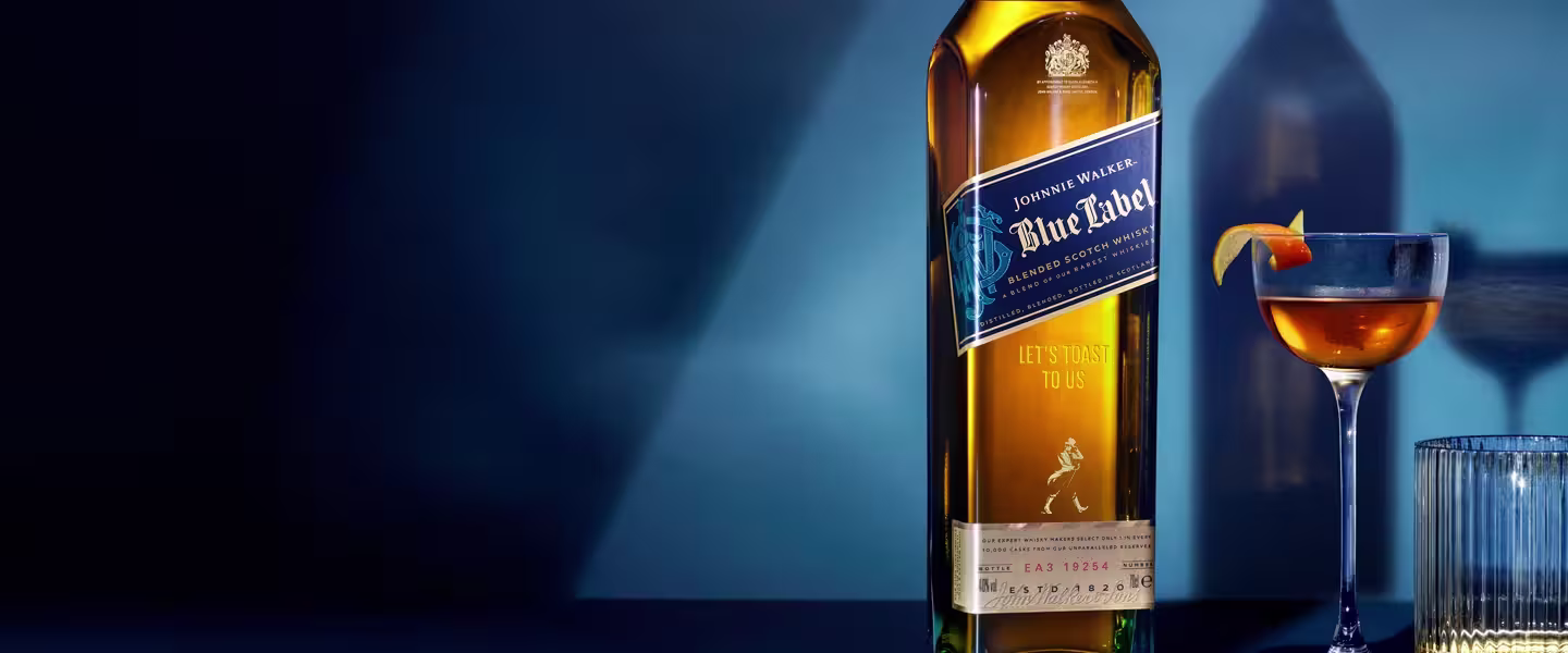 Tall bottle of Johnnie Walker Blue Label whisky beside a cocktail glass with an orange twist garnish and a glass of water, set against a blue background with dramatic lighting.